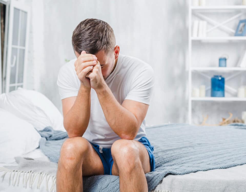 Imagem de um homem sentado na cama com as mãos na cabeça para simbolizar dor nos testículos após a relação sexual