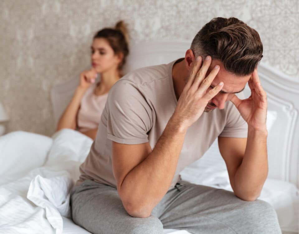 Imagem de um homem sentado na cama com as mãos na cabeça e a mulher atrás, ambos preocupados e tristes, simbolizando a azoospermia