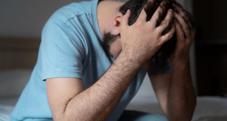 Imagem de um homem sentado na cama com as mãos na cabeça com a expressão preocupada para simbolizar o pênis ressecado