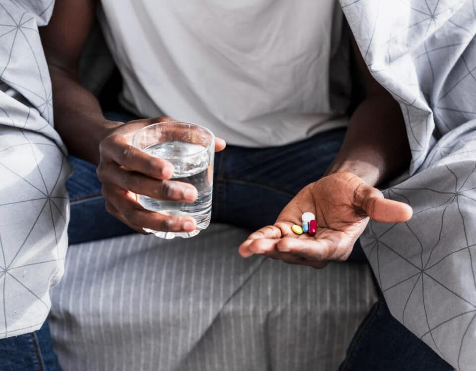 Imagem de um homem sentado na cama com remédios numa mão e um copo de água na outra para simbolizar os remédios psiquiátricos