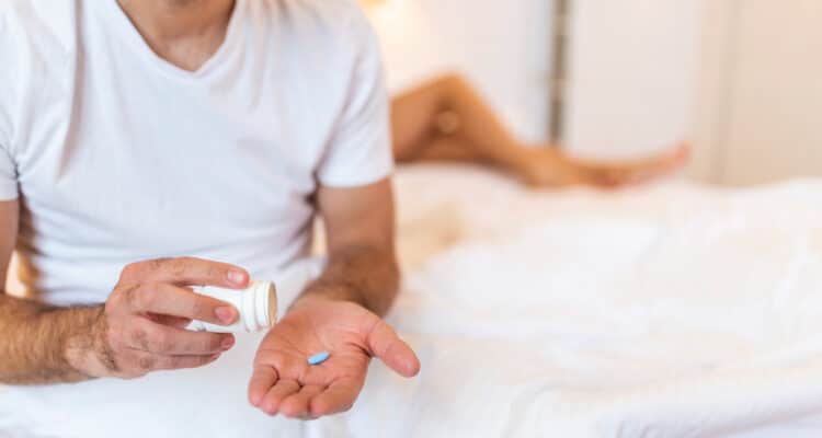 Imagem de um homem com um comprimido na mão e os pés de uma mulher na cama atrás dele para simbolizar o chemsex