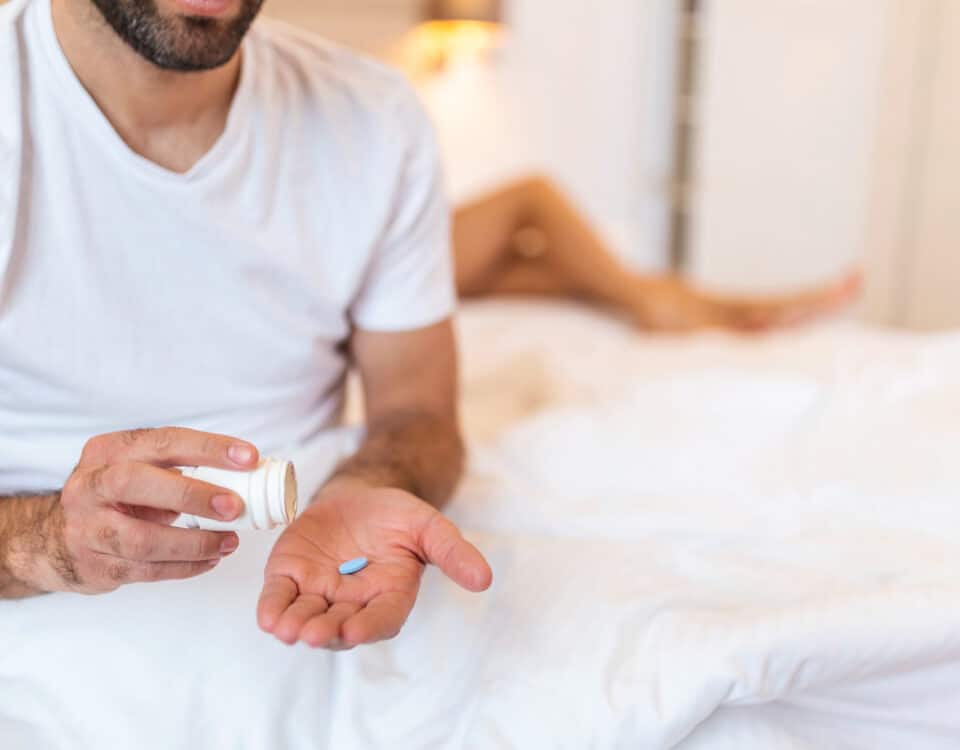 Imagem de um homem com um comprimido na mão e os pés de uma mulher na cama atrás dele para simbolizar o chemsex