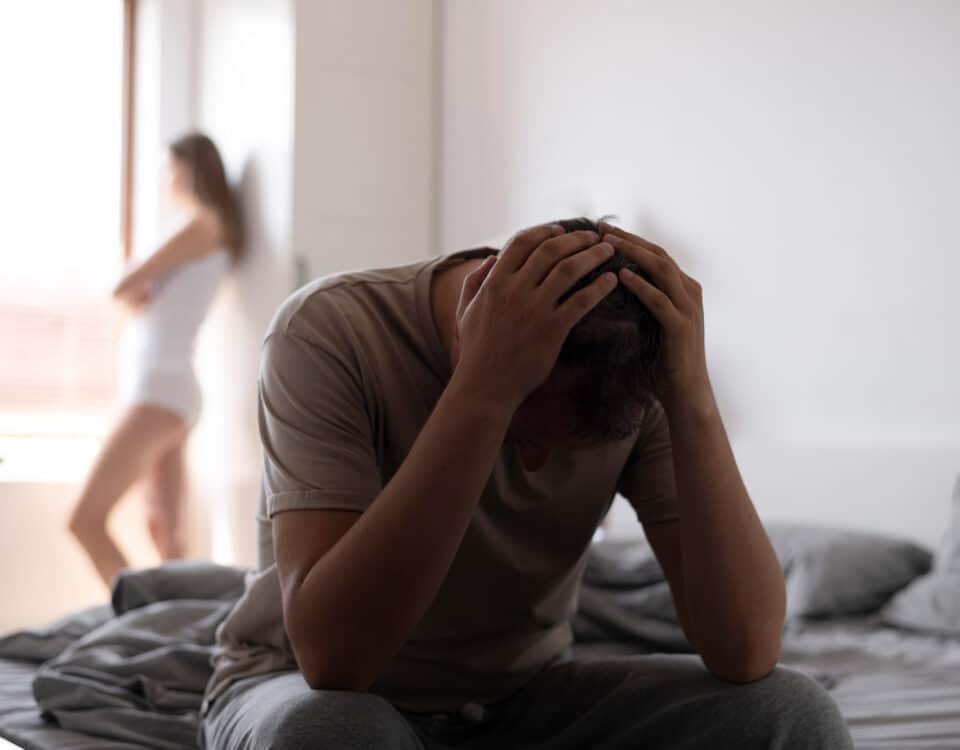 Imagem de um homem na cama com as mãos na cabeça e a cabeça pra baixo e uma mulher na janela olhando pro nada, para simbolizar a injeção peniana